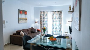 A living room with a brown sofa, a glass dining table with chairs, and a floral curtain. - Cosy APT in Sliema- 2 mins walk from sea (Sliema)