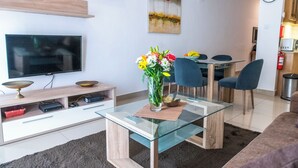 A modern living room with a glass coffee table, a flat-screen TV, a dining area with a glass table, and a painting on the wall. - Cosy APT in Sliema- 2 mins walk from sea (Sliema)