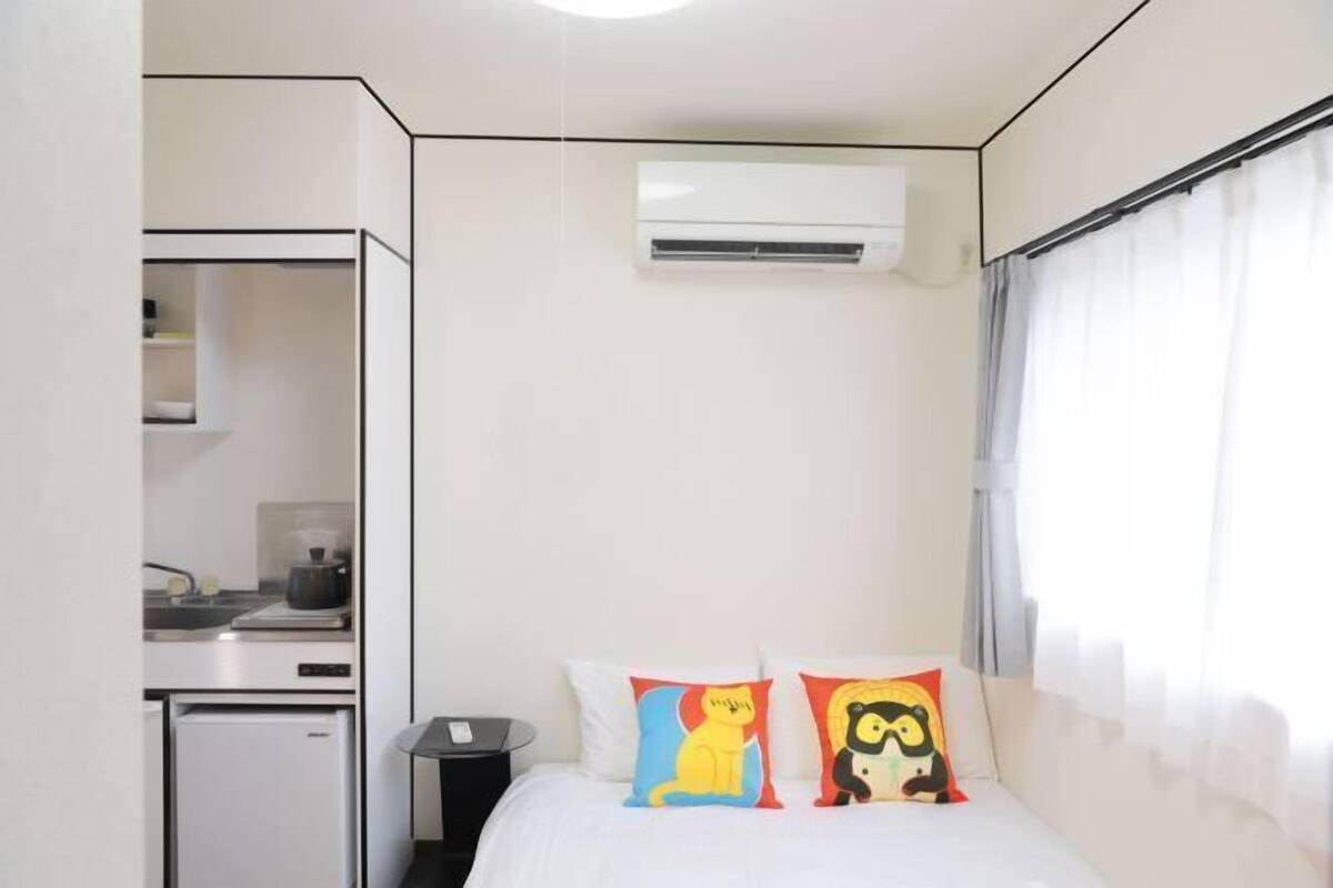 A compact hotel room with a kitchenette, a bed with colorful pillows, and an air conditioning unit.