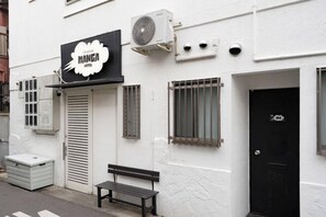 A building with a sign that reads 'MANGA HOTEL,' a black door, a bench, and an air conditioning unit. - Oyasumi manga hotel (Osaka)