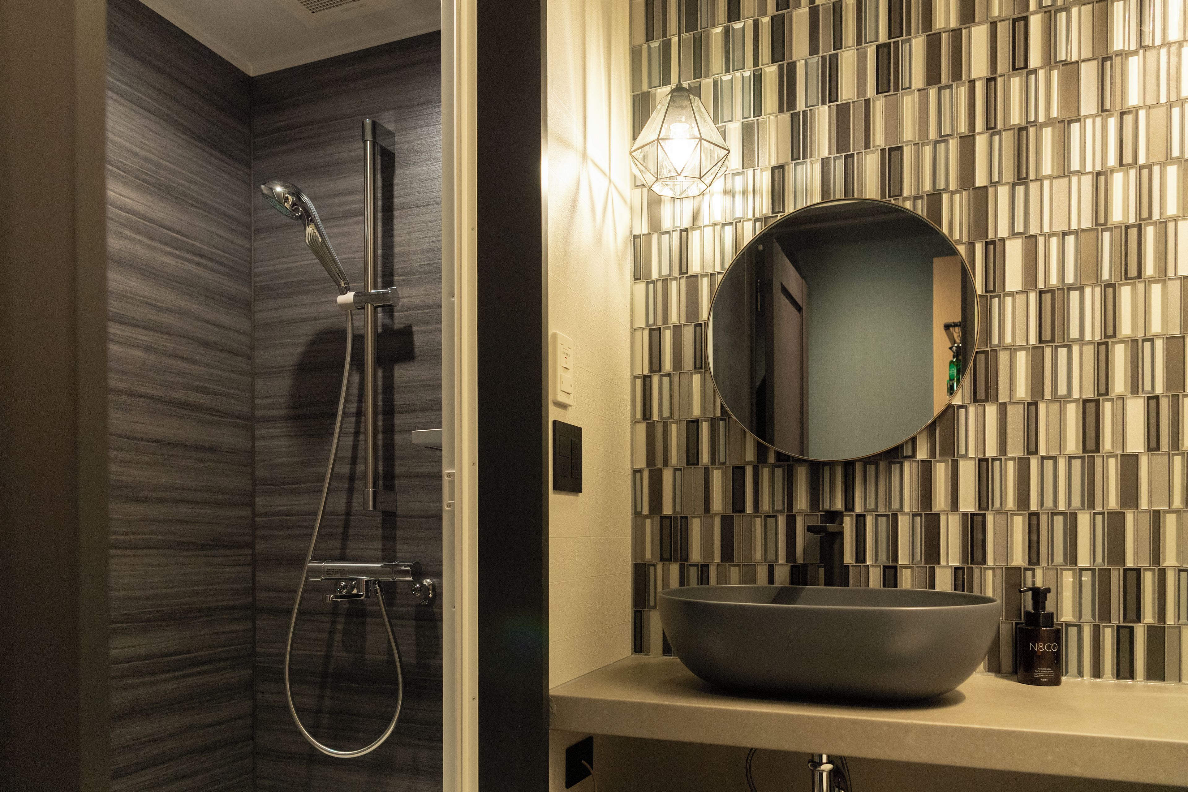 A bathroom with a modern design, featuring a shower with a handheld showerhead and a wall-mounted faucet, a round mirror above a sink with a vessel basin, and a patterned wall.
