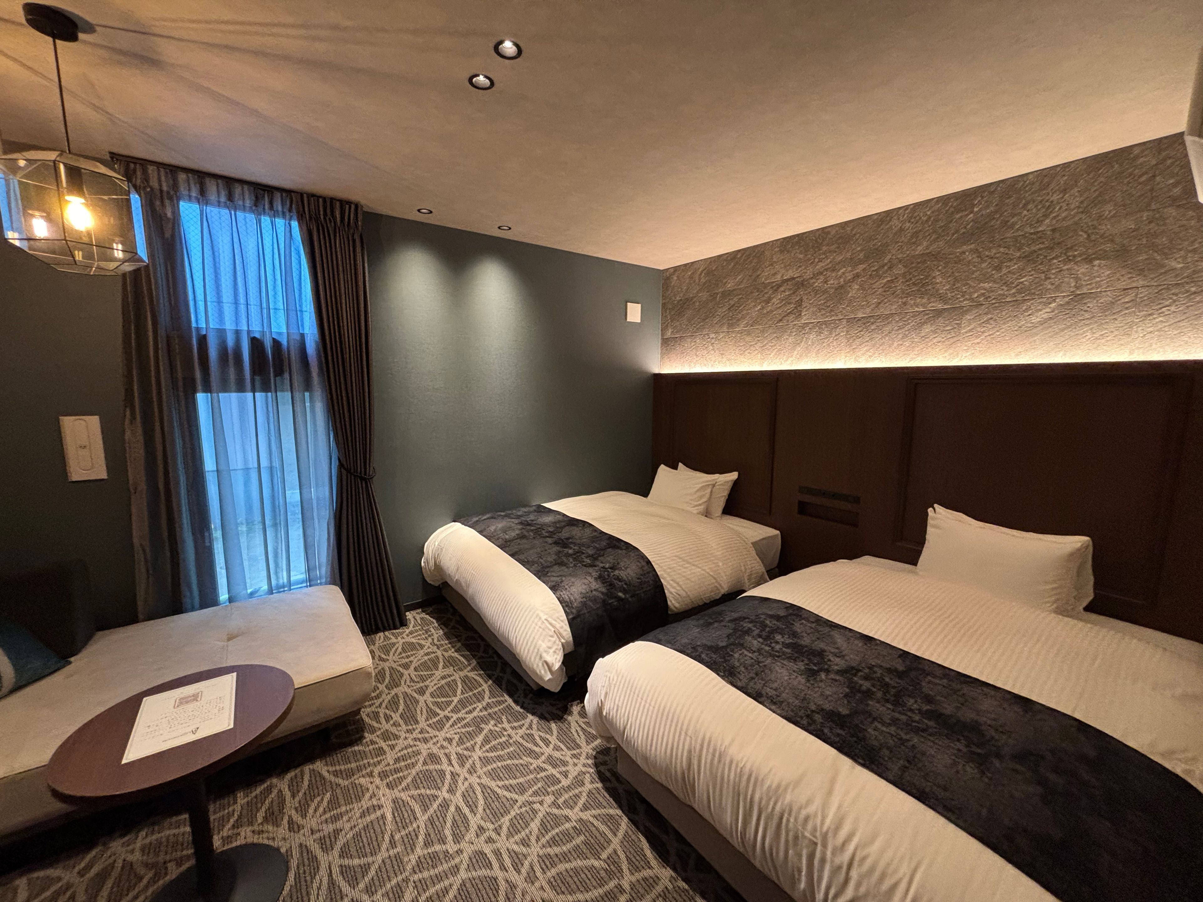 A hotel room with two beds, a small table, and a window with curtains.