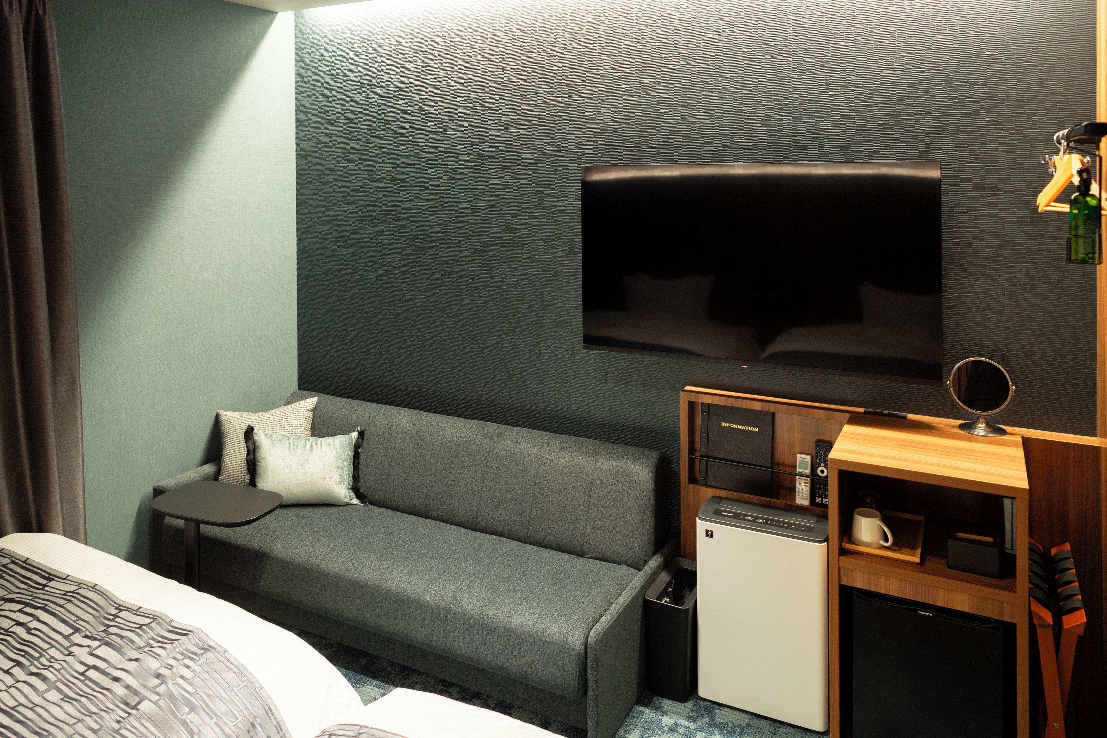 A hotel room with a grey sofa, a small bedside table, a TV mounted on the wall, and a compact kitchenette.