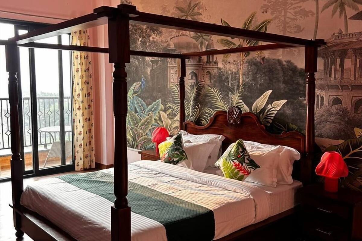A four-poster bed with a canopy, a nightstand with a lamp, and a mural of a tropical landscape on the wall.