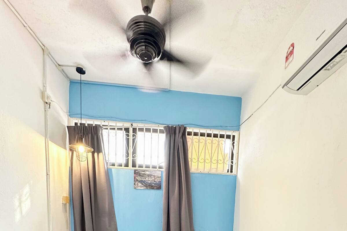 A small, single-room hotel room with two beds, a ceiling fan, a small table with a lamp, and a window with curtains.