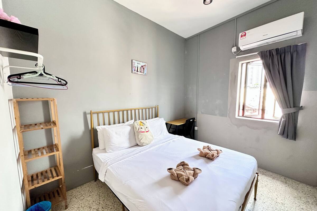 A bedroom with a bed, a wooden shelf, a hanging rack, a window with curtains, and an air conditioning unit.