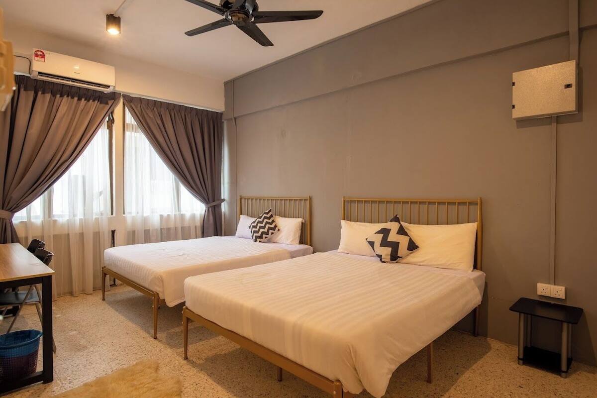 A hotel room with two single beds, a ceiling fan, an air conditioning unit, a small table, and a desk.