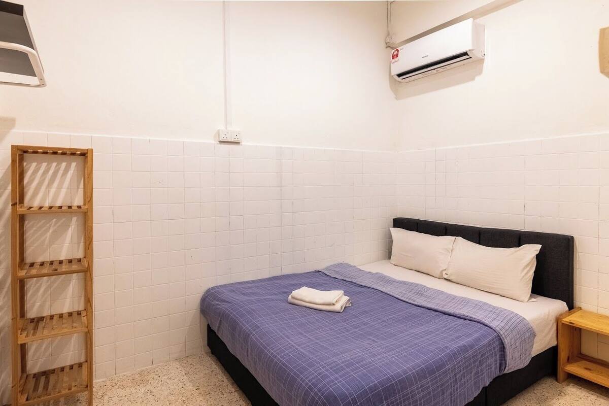 A small, neatly arranged bedroom with a bed, a wooden shelf unit, and a wall-mounted air conditioner.
