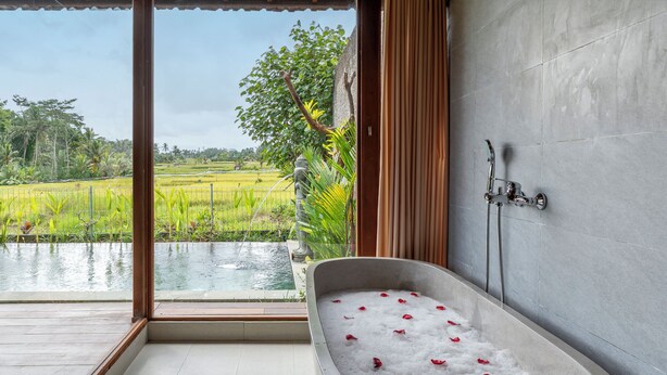 Een badkamer met een bad vol water en rozenblaadjes, een douche en uitzicht op een rijstveld.