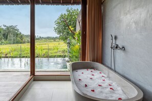 Un baño con una bañera llena de agua y pétalos de rosa, ducha y vistas a un campo de arroz.