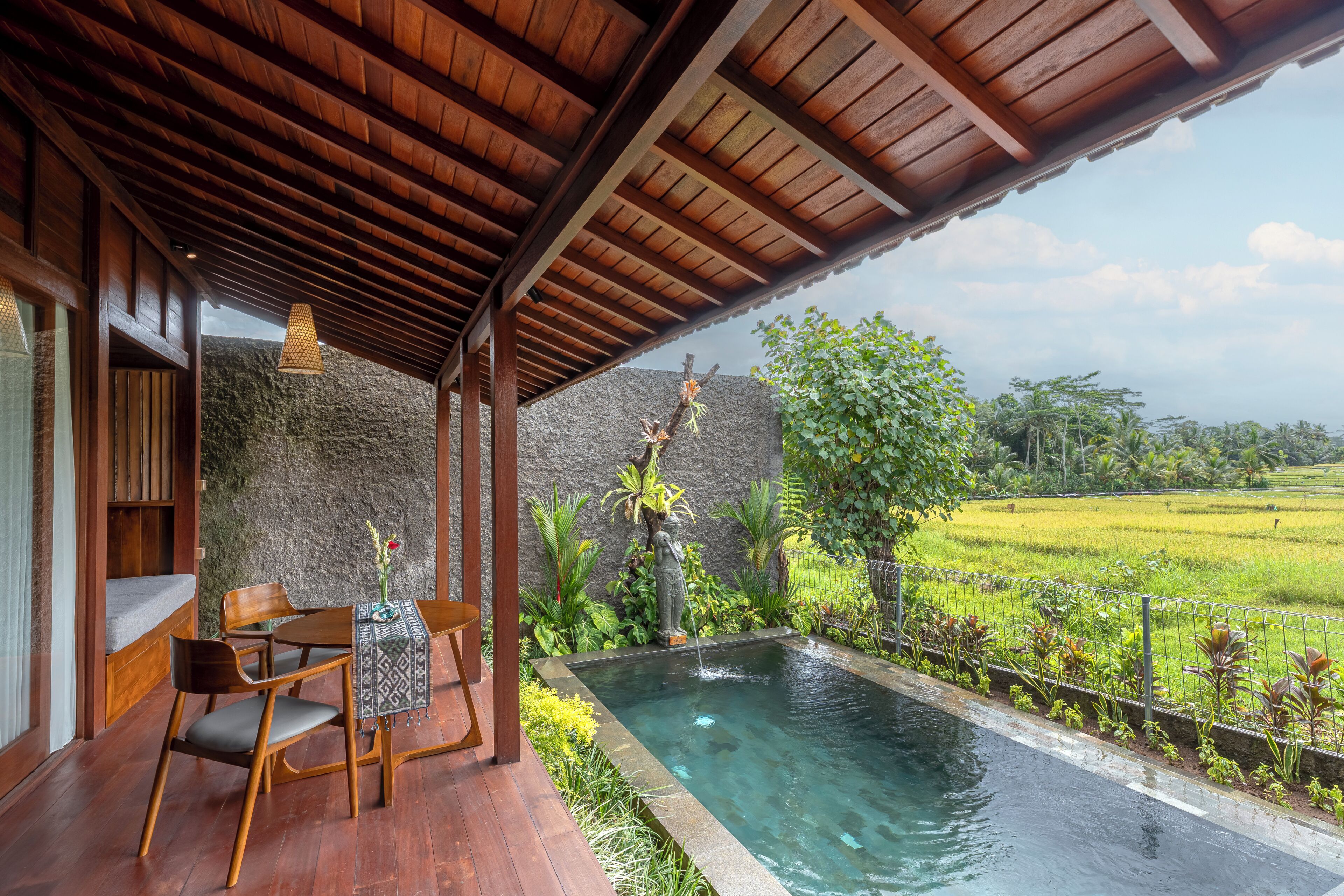 Una terraza de madera con piscina, un muro de piedra y vistas a un campo de arroz.
