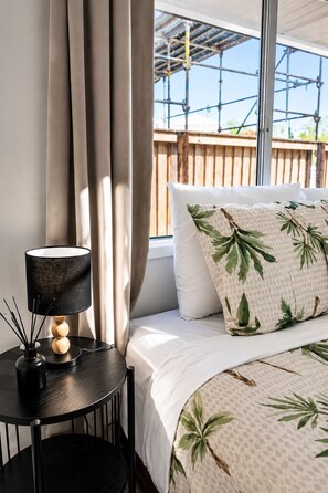 A bedroom with a bed, a bedside table with a lamp, a window with curtains, and a view of a wooden deck.