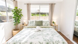 Ein Schlafzimmer mit einem großen Bett, einer Zimmerpflanze, zwei Nachttischen mit Lampen und Blick durch ein Fenster.