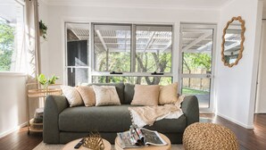 Ein modernes Wohnzimmer mit einem grauen Sofa, zwei runden Holztischen, einem gewobenen Pouf und einem großen Fenster mit Blick auf Grünflächen.