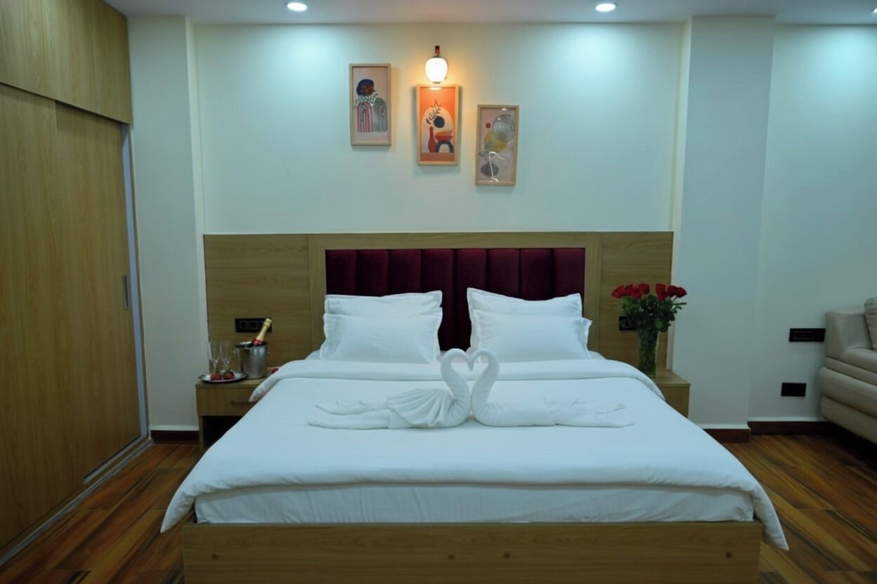 A neatly made bed with white linens and a heart-shaped towel arrangement, a wooden headboard, a nightstand with a vase of red flowers, and a wall-mounted light fixture.