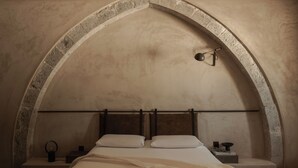 Une chambre à coucher avec un plafond en forme d’arc en pierre, un lit recouvert de draps blancs, une table de chevet avec une lampe et une petite table de nuit dotée d’un anneau lumineux.