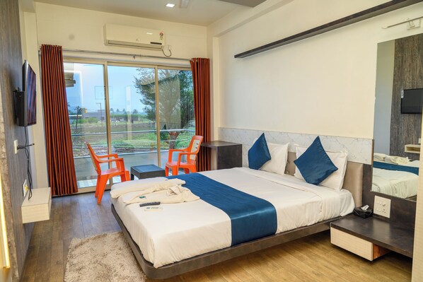 A modern hotel room with a large bed, a flat-screen TV, a balcony with orange chairs, and a large window. - Hotel Srushti Regency (Wai)