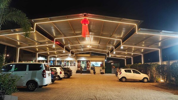 A well-lit, modern building with a covered parking area and several cars parked in front. - Hotel Srushti Regency (Wai)