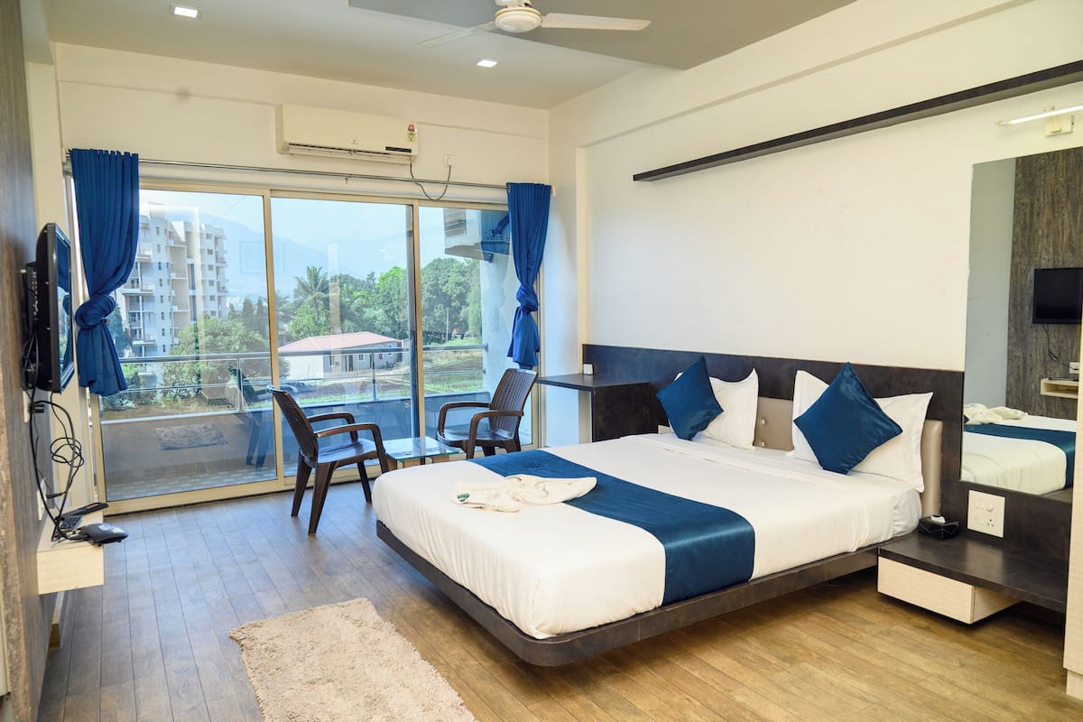 A modern hotel room with a large bed, a desk, and a chair. There is a view of buildings and trees through the window.