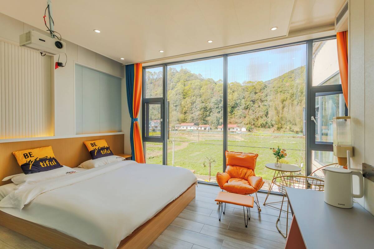 A modern bedroom with a large bed, a view of a green landscape, a comfortable orange chair, and a small table with a lamp.