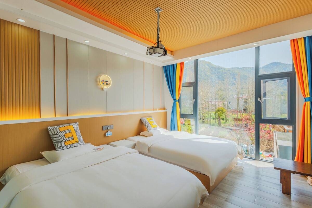 A modern hotel room with two beds, a large window with curtains, a ceiling projector, and a view of the outdoors.