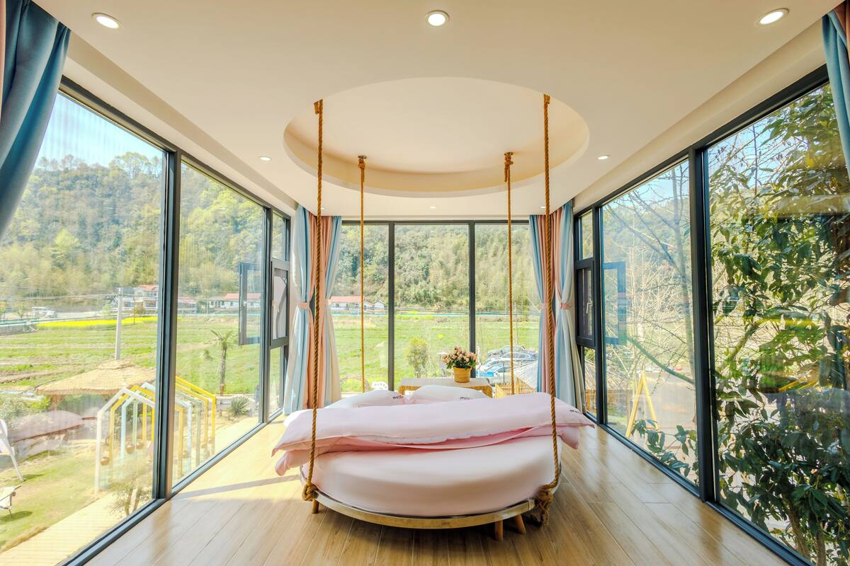 A modern bedroom with a swing bed, large windows, and a view of a green landscape.