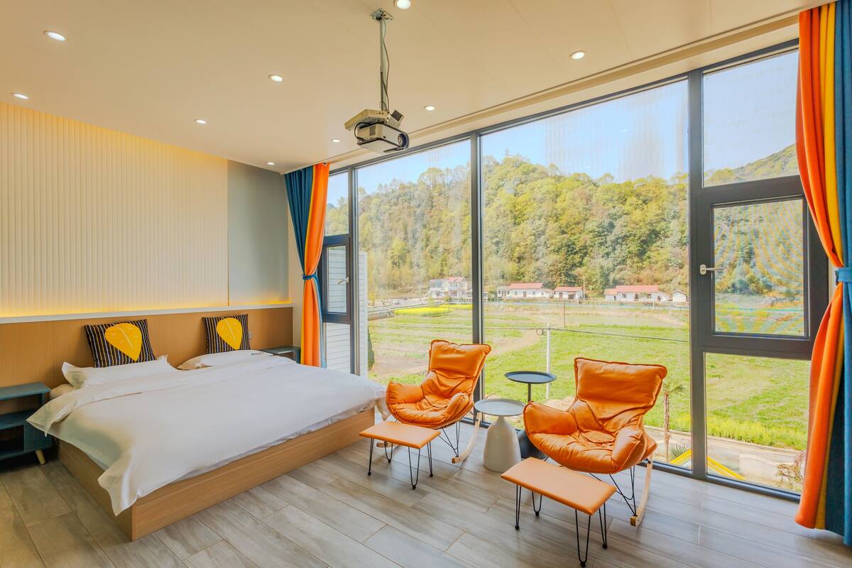 A modern bedroom with a large bed, two orange leather chairs, a small table, and a view of a green landscape through large windows.