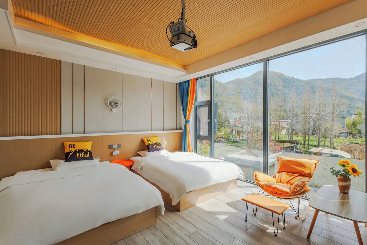 A modern hotel room with two beds, a large window with a mountain view, a wooden ceiling, and a small table with a vase.