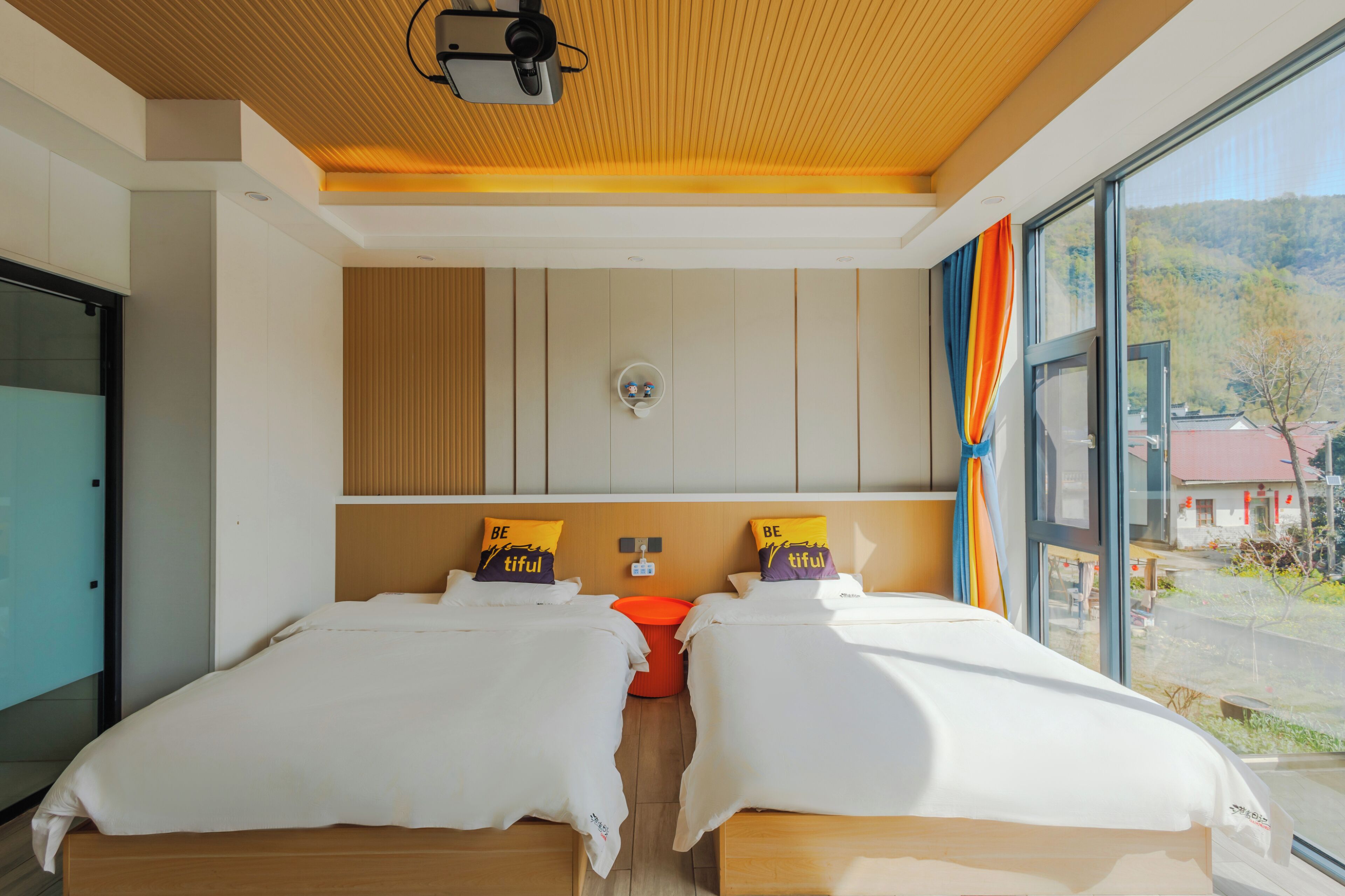 Una habitación de hotel moderna con dos camas, techo de madera, un ventanal con vistas al exterior y una pequeña mesa con un cubo rojo.