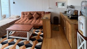 Un espacio habitable compacto con sofá, una pequeña mesa de madera, una zona de cocina con dispensador de agua y un ventanal que proporciona luz natural.