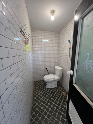 Une salle de bain avec des murs recouverts de carreaux blancs, des toilettes, un espace douche et une porte dotée d’un panneau en verre.
