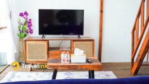 Una sala de estar con un televisor sobre un mueble de madera, una mesita de mimbre con botellas y una caja de pañuelos, y una escalera a la derecha.