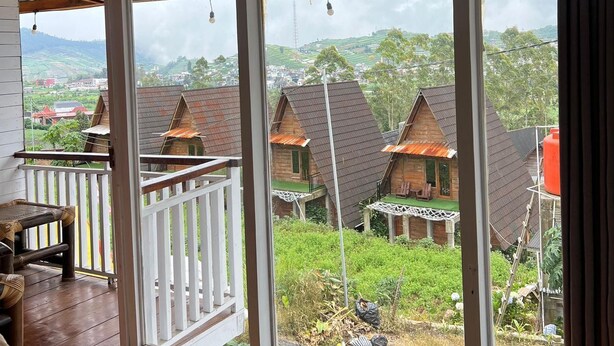 Una habitación con un gran ventanal corredero que da a un balcón. La habitación tiene piso de madera y barandilla blanca en el balcón.