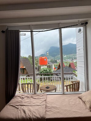 Une chambre avec un lit, un balcon doté d’une table et de chaises, et une vue sur un village avec ses maisons et une montagne.