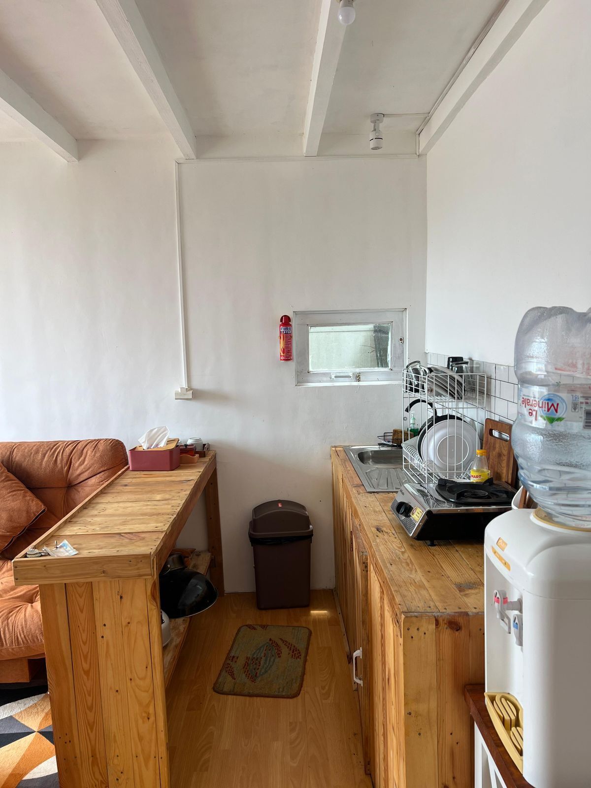 Cocina compacta con encimera de madera, dispensador de agua, escurreplatos y un pequeño cubo de basura.