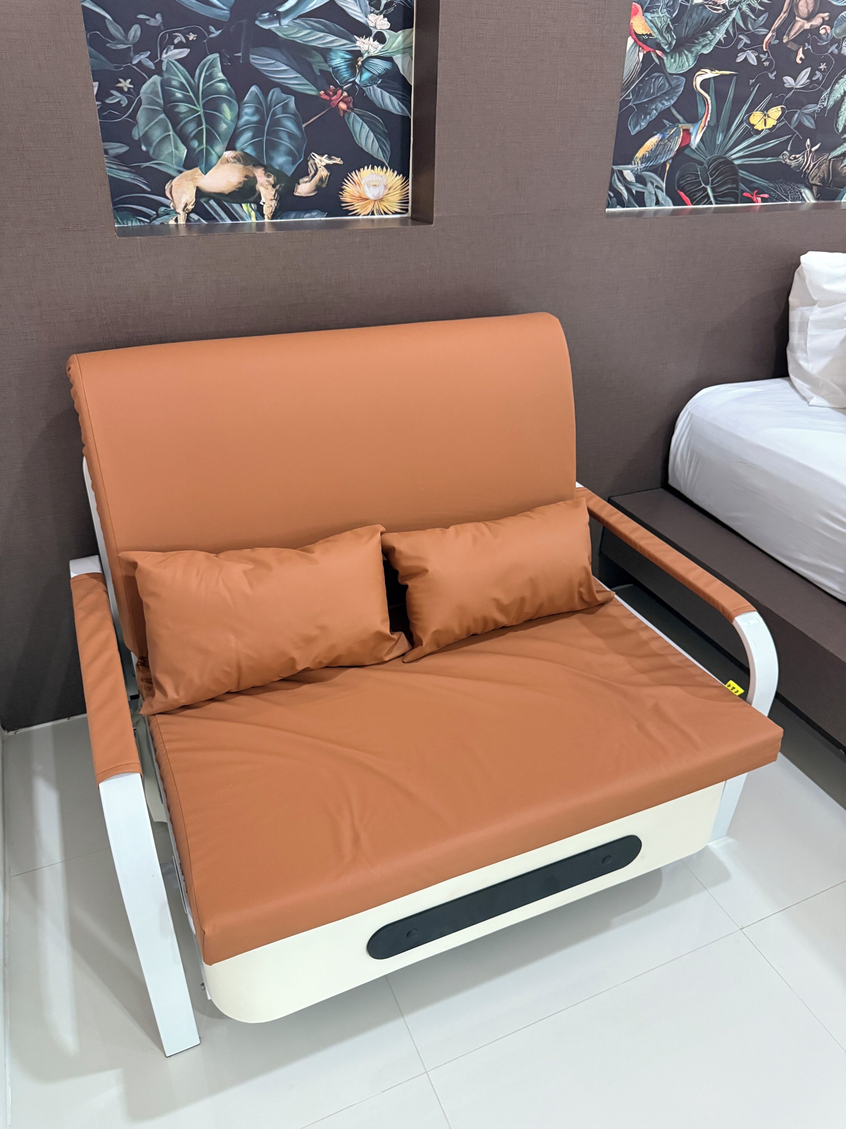 Um sofá moderno com uma almofada marrom em um quarto com parede cinza e uma cama branca.