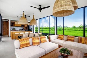 A modern living room with a sofa set, a coffee table, and a view of a green field through large windows. - Villa Samaddhi by BaliSuperHost (Bali)