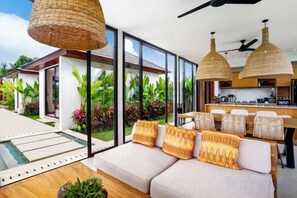 A modern living room with a sofa, a dining area, and a kitchen with large windows. - Villa Samaddhi by BaliSuperHost (Bali)