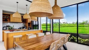 A modern kitchen with a central island, large windows, and a dining area with a wooden table and wicker chairs. - Villa Samaddhi by BaliSuperHost (Bali)