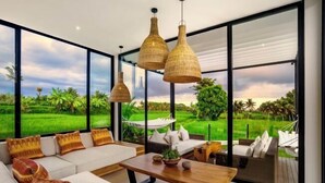 A modern living room with a large glass wall, a sofa set, a wooden coffee table, and hanging wicker lamps. - Villa Samaddhi by BaliSuperHost (Bali)