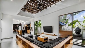 A modern living room with a long wooden bench, a flat-screen TV, and a large window with a view of greenery. - Villa Lina by BaliSuperHost (Bali)