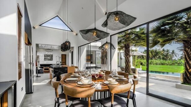 A modern dining area with a round table set for four, surrounded by chairs with wooden legs and black cushions. The room features large windows offering a view of a lush garden and a swimming pool. - Villa Lina by BaliSuperHost (Bali)