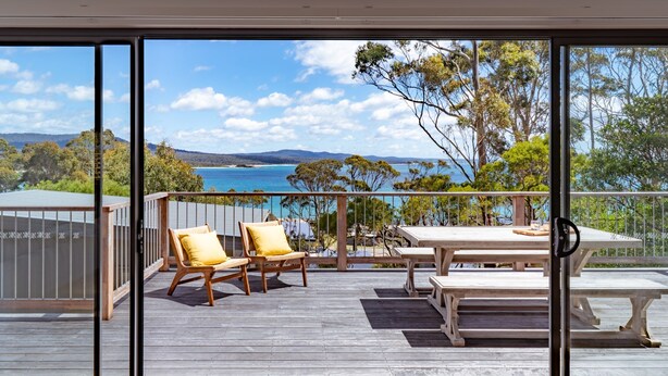 A balcony with wooden furniture, a table, and chairs, overlooking a scenic landscape with trees and a body of water. - Marmoris Bay of Fires - A laid back coastal retreat with shimmering ocean views (Binalong Bay)