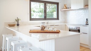 A modern kitchen with white cabinetry, a large island with stools, a wooden cutting board, and a window with a view of trees. - Marmoris Bay of Fires - A laid back coastal retreat with shimmering ocean views (Binalong Bay)