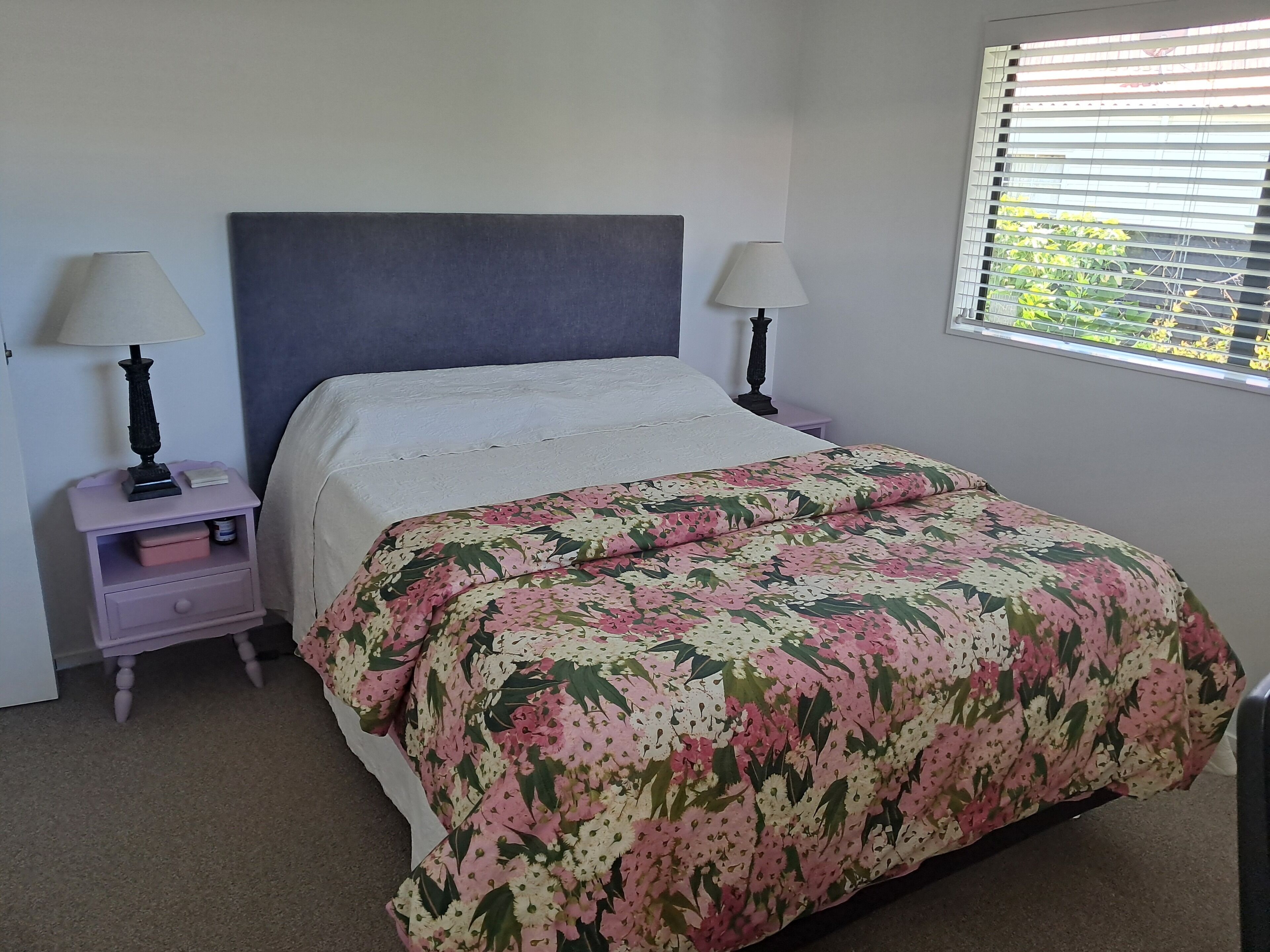 A bedroom with a bed, two bedside tables with lamps, a nightstand, and a window with blinds.