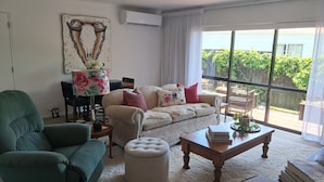 Un salon comprenant un canapé, un pouf, une table basse et un fauteuil. Une grande fenêtre offre une vue sur la verdure à l’extérieur.