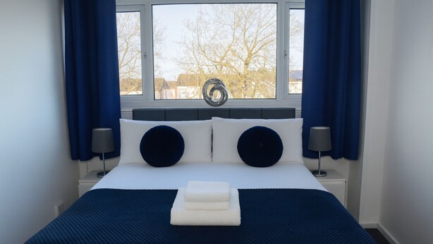A neatly made bed with a blue and white color scheme, a window with a view of trees, and two bedside lamps. - Stylish 3-Bed House in Wolverhampton | Long Stays Welcome | Near Hospital (West Midlands)