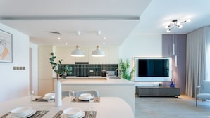 Een moderne keuken met een centraal eiland, een eetgedeelte met een witte tafel voor vier personen, en een zithoek met een grote flatscreen-tv en een grijze bank.