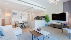 Een moderne woonkamer met een bank, een salontafel en een televisie. Er is een eethoek met stoelen en een tafel, en een keuken op de achtergrond.