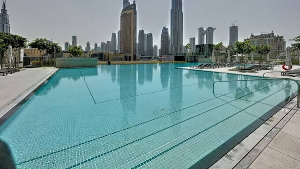 Una piscina con agua azul cristalina, rodeada por una solana con baldosas. Al fondo, varios edificios altos, incluido un rascacielos muy alto.